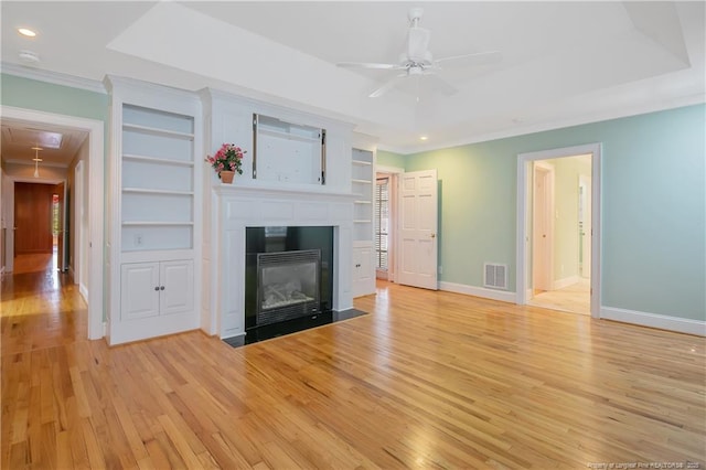 unfurnished living room featuring built in features, ceiling fan, a tray ceiling, ornamental molding, and light hardwood / wood-style floors