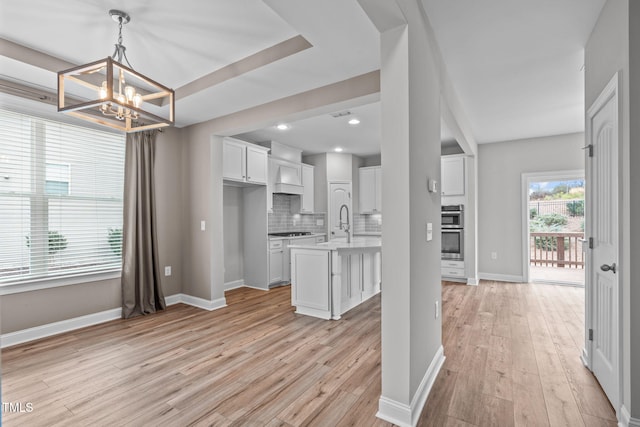 kitchen with white cabinetry, hanging light fixtures, light hardwood / wood-style floors, a kitchen island with sink, and backsplash