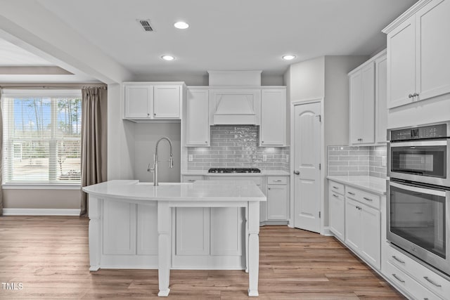 kitchen with black gas cooktop, white cabinetry, custom range hood, stainless steel double oven, and light wood-type flooring