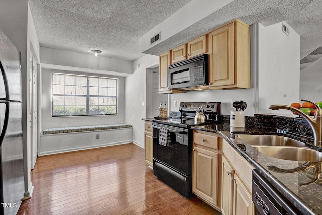 kitchen with light brown cabinetry, sink, light hardwood / wood-style flooring, dark stone countertops, and black appliances