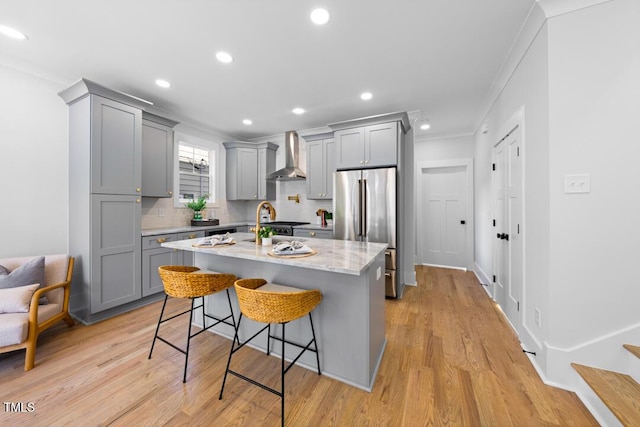 kitchen featuring gray cabinets, a breakfast bar, stainless steel refrigerator, an island with sink, and wall chimney range hood