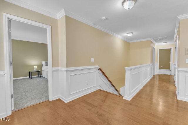 hallway featuring light wood finished floors, visible vents, crown molding, an upstairs landing, and wainscoting