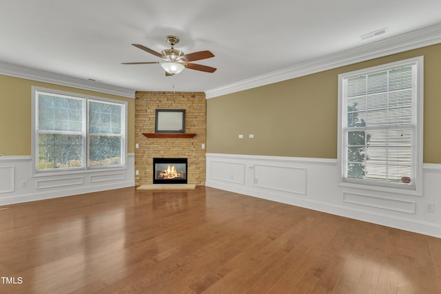 unfurnished living room with visible vents, wood finished floors, a stone fireplace, crown molding, and ceiling fan