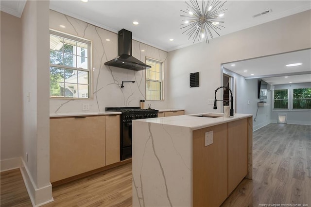 kitchen with black range with gas stovetop, wall chimney exhaust hood, an island with sink, and light wood-type flooring