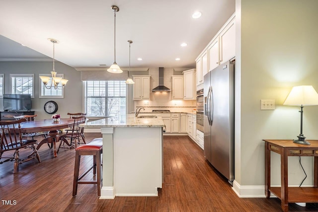 kitchen with wall chimney range hood, stainless steel fridge, a kitchen island with sink, light stone counters, and decorative light fixtures