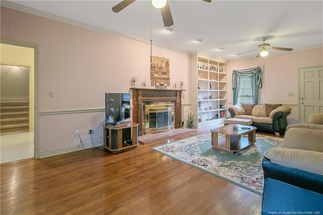 living room featuring built in shelves, crown molding, ceiling fan, a fireplace, and hardwood / wood-style floors