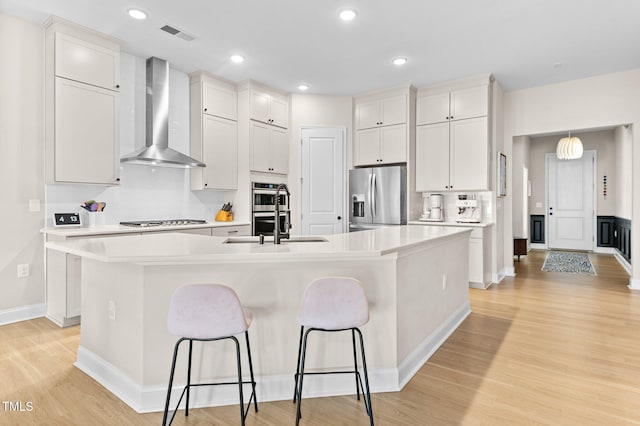 kitchen featuring wall chimney exhaust hood, white cabinetry, appliances with stainless steel finishes, and an island with sink