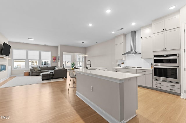 kitchen with wall chimney exhaust hood, light hardwood / wood-style flooring, a kitchen breakfast bar, stainless steel appliances, and a kitchen island with sink