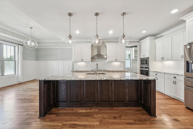 kitchen with white cabinetry, decorative light fixtures, a center island with sink, and wall chimney range hood