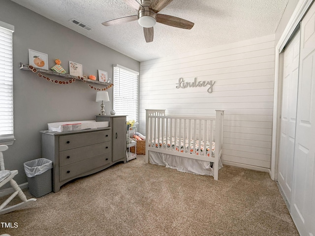 bedroom with a textured ceiling, ceiling fan, a closet, a nursery area, and light colored carpet