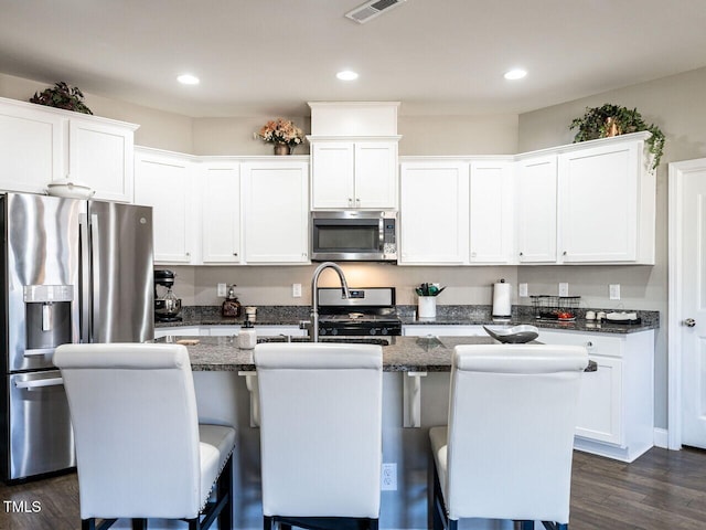 kitchen featuring white cabinetry, appliances with stainless steel finishes, a center island with sink, and dark stone countertops