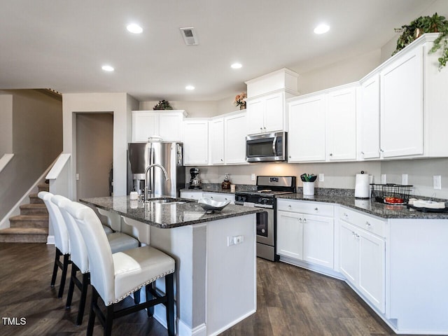 kitchen featuring appliances with stainless steel finishes, white cabinets, dark stone counters, dark wood-type flooring, and a center island with sink