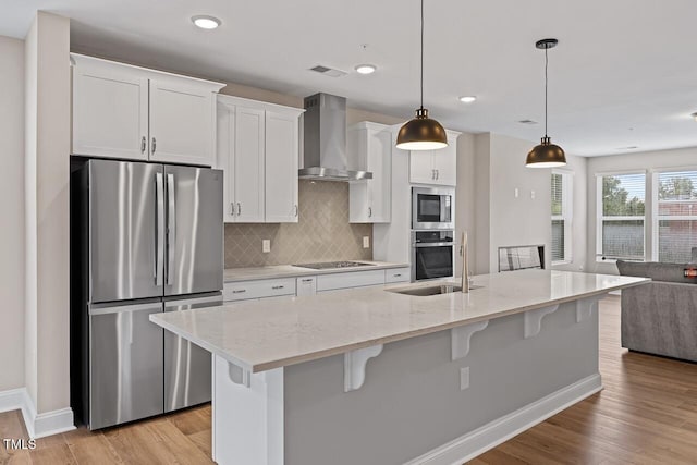 kitchen with wall chimney exhaust hood, white cabinetry, an island with sink, pendant lighting, and stainless steel appliances