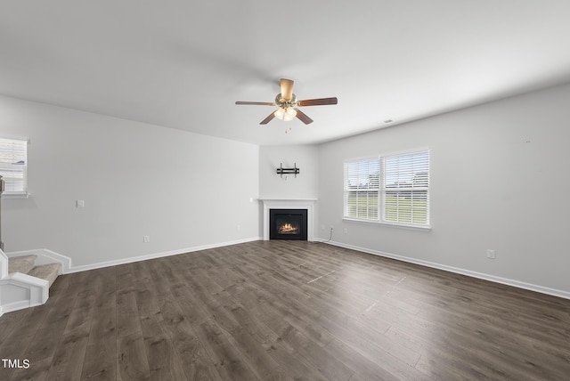 unfurnished living room featuring a ceiling fan, dark wood-style flooring, a glass covered fireplace, baseboards, and stairs