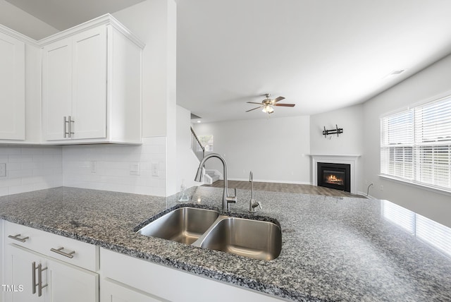 kitchen with a sink, a glass covered fireplace, white cabinetry, dark stone counters, and decorative backsplash