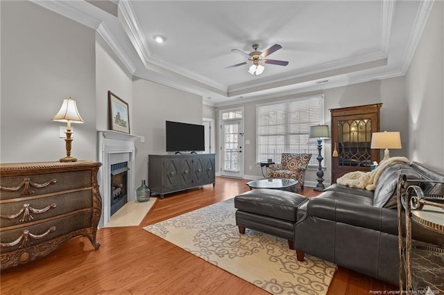 living area with ornamental molding, a fireplace with flush hearth, a raised ceiling, ceiling fan, and wood finished floors