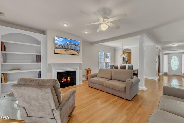 living area with a lit fireplace, light wood finished floors, decorative columns, and a ceiling fan