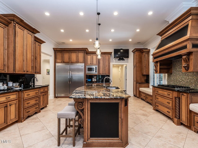 kitchen with sink, built in appliances, an island with sink, decorative light fixtures, and dark stone counters