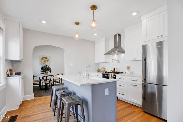 kitchen featuring wall chimney exhaust hood, white cabinetry, hanging light fixtures, stainless steel fridge, and a kitchen island with sink