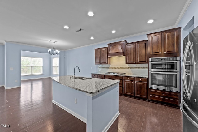 kitchen featuring sink, stainless steel appliances, light stone countertops, custom range hood, and an island with sink