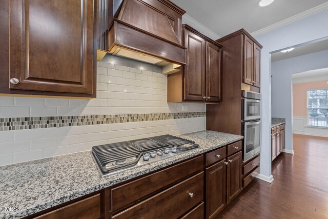 kitchen featuring dark brown cabinetry, custom exhaust hood, ornamental molding, stainless steel appliances, and light stone countertops