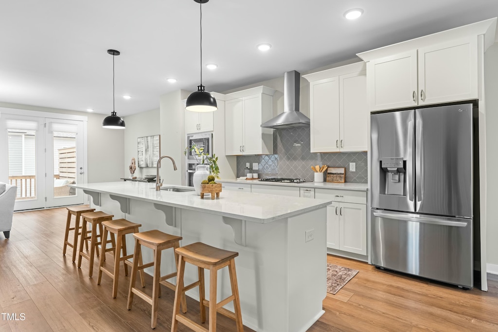 kitchen featuring wall chimney exhaust hood, decorative light fixtures, appliances with stainless steel finishes, a kitchen island with sink, and white cabinets
