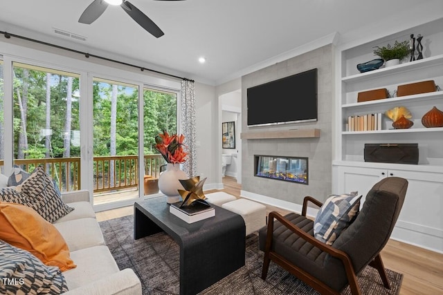 living room featuring built in features, ceiling fan, ornamental molding, a large fireplace, and light wood-type flooring