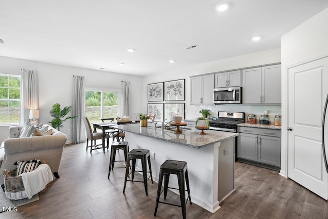 kitchen with gray cabinets, appliances with stainless steel finishes, a breakfast bar, light stone countertops, and a center island with sink