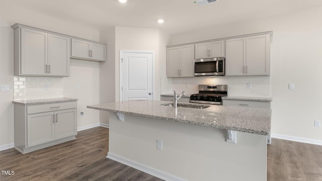 kitchen featuring sink, appliances with stainless steel finishes, wood-type flooring, light stone countertops, and a center island with sink