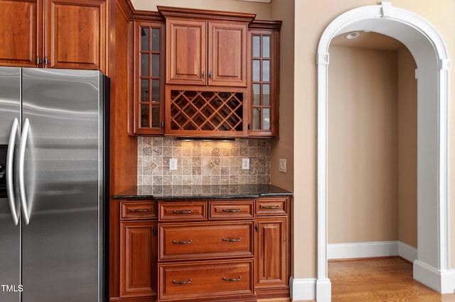 bar with light wood-type flooring, decorative backsplash, dark stone counters, and stainless steel fridge with ice dispenser