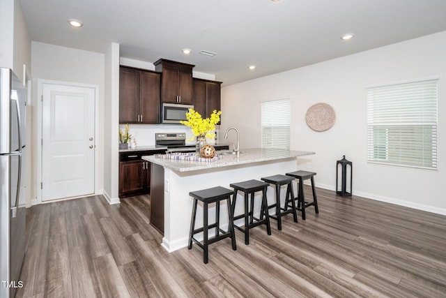 kitchen featuring appliances with stainless steel finishes, dark hardwood / wood-style flooring, a kitchen bar, dark brown cabinetry, and a center island with sink