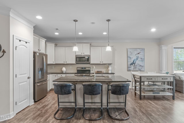 kitchen featuring a kitchen island with sink, a sink, hanging light fixtures, appliances with stainless steel finishes, and dark stone counters