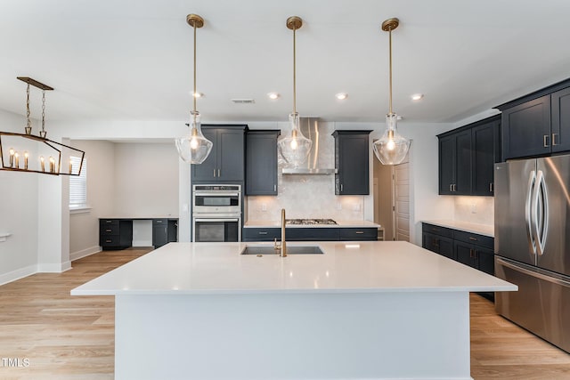 kitchen with pendant lighting, wall chimney range hood, sink, appliances with stainless steel finishes, and an island with sink