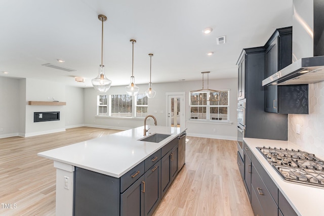 kitchen featuring wall chimney exhaust hood, sink, pendant lighting, stainless steel appliances, and a kitchen island with sink