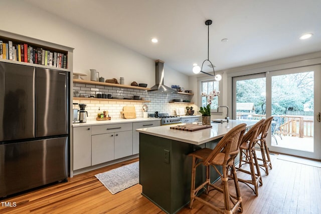 kitchen with stainless steel refrigerator, white cabinetry, a center island with sink, decorative light fixtures, and wall chimney exhaust hood