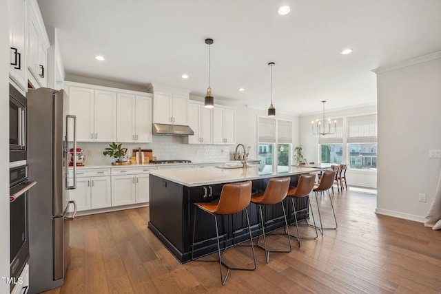 kitchen with appliances with stainless steel finishes, pendant lighting, tasteful backsplash, white cabinets, and a center island with sink