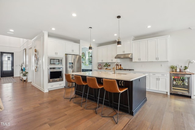 kitchen with appliances with stainless steel finishes, white cabinetry, hanging light fixtures, wine cooler, and an island with sink