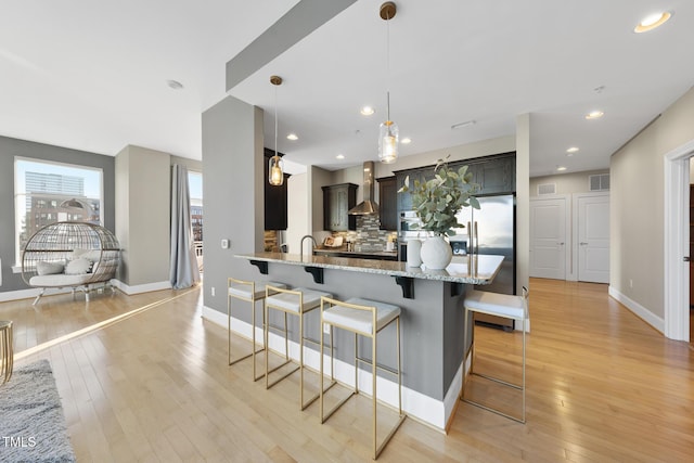 kitchen featuring wall chimney range hood, tasteful backsplash, light stone countertops, a kitchen bar, and kitchen peninsula