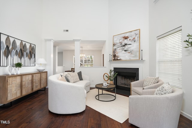 living area featuring a fireplace with flush hearth, baseboards, a towering ceiling, decorative columns, and dark wood finished floors