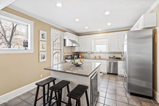 kitchen with appliances with stainless steel finishes, white cabinetry, a kitchen island, stone countertops, and wall chimney exhaust hood