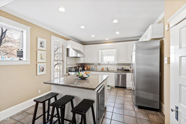 kitchen with a kitchen island, stainless steel appliances, a kitchen bar, exhaust hood, and dark stone counters