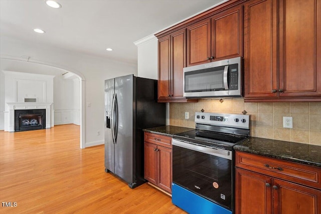 kitchen with arched walkways, stainless steel appliances, light wood-type flooring, decorative backsplash, and dark stone countertops