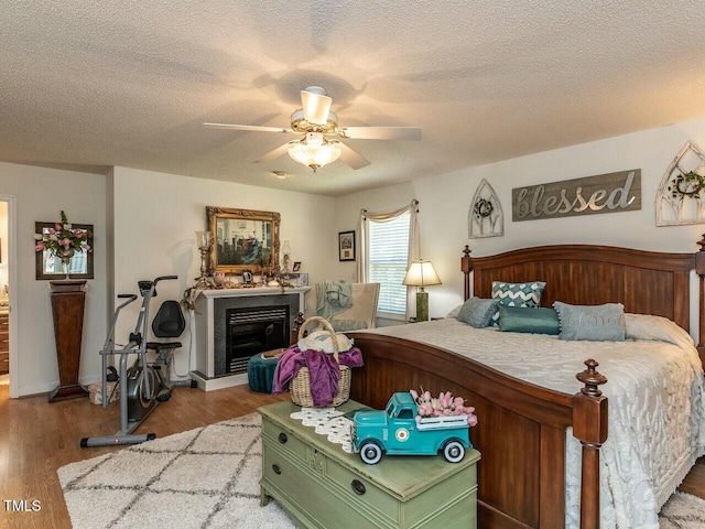 bedroom with a ceiling fan, a textured ceiling, wood finished floors, and a glass covered fireplace