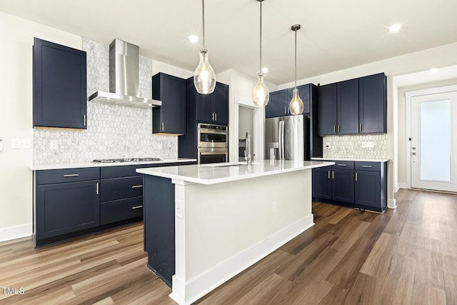 kitchen featuring wall chimney range hood, a kitchen island with sink, hanging light fixtures, stainless steel appliances, and dark hardwood / wood-style flooring