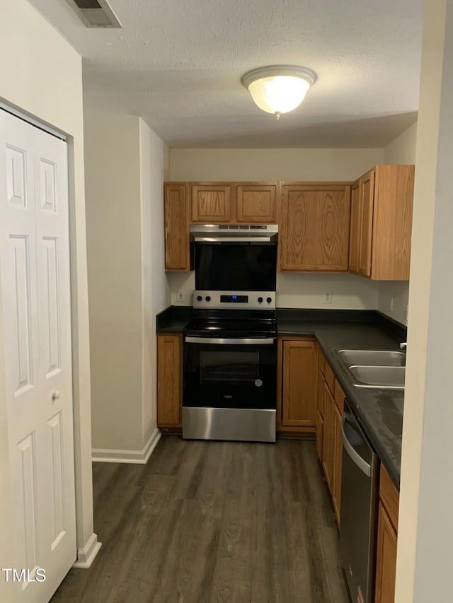 kitchen with under cabinet range hood, a sink, appliances with stainless steel finishes, dark wood-style floors, and dark countertops