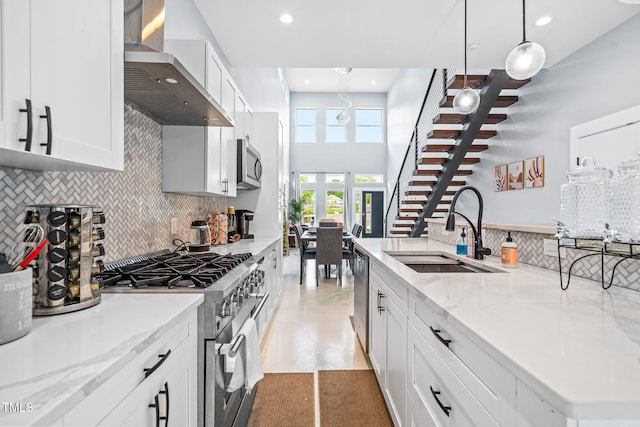 kitchen featuring wall chimney exhaust hood, light stone counters, hanging light fixtures, stainless steel appliances, and a sink