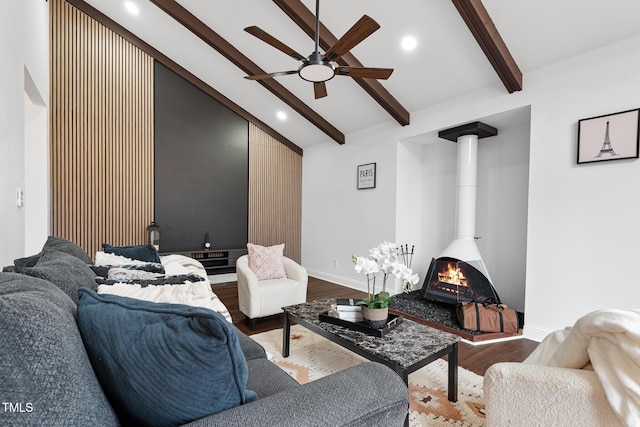 living room featuring beam ceiling, wood-type flooring, ceiling fan, and a wood stove