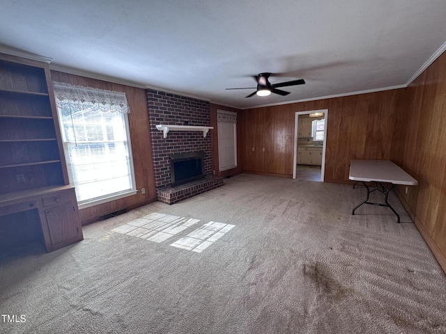 unfurnished living room with a fireplace, wooden walls, ceiling fan, crown molding, and light carpet