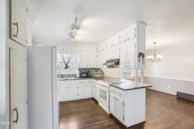 kitchen with dark countertops, under cabinet range hood, a wainscoted wall, white appliances, and a sink
