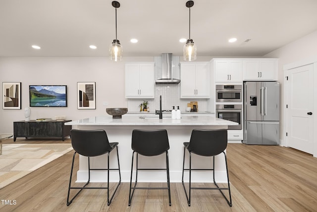 kitchen featuring wall chimney exhaust hood, light stone counters, an island with sink, stainless steel appliances, and white cabinets